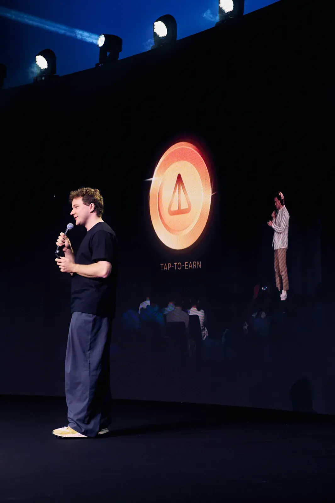 Speaker with microphone on stage at a Dubai investment event, with audience and TAP-TO-EARN screen behind